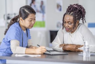 A senor patient of African decent, sits across the desk from her female doctor as they go over her recent lab results on a tablet in front of them.  The woman is dressed casually and leaning in closely to look at the screen as the doctor explains what she is seeing.