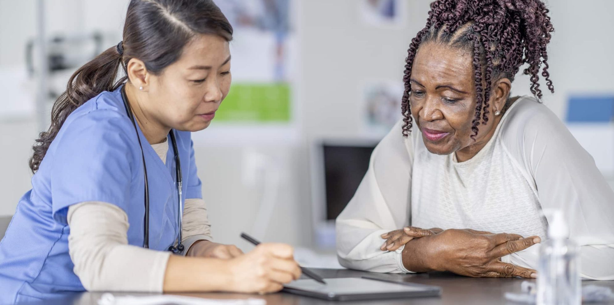 A senor patient of African decent, sits across the desk from her female doctor as they go over her recent lab results on a tablet in front of them.  The woman is dressed casually and leaning in closely to look at the screen as the doctor explains what she is seeing.